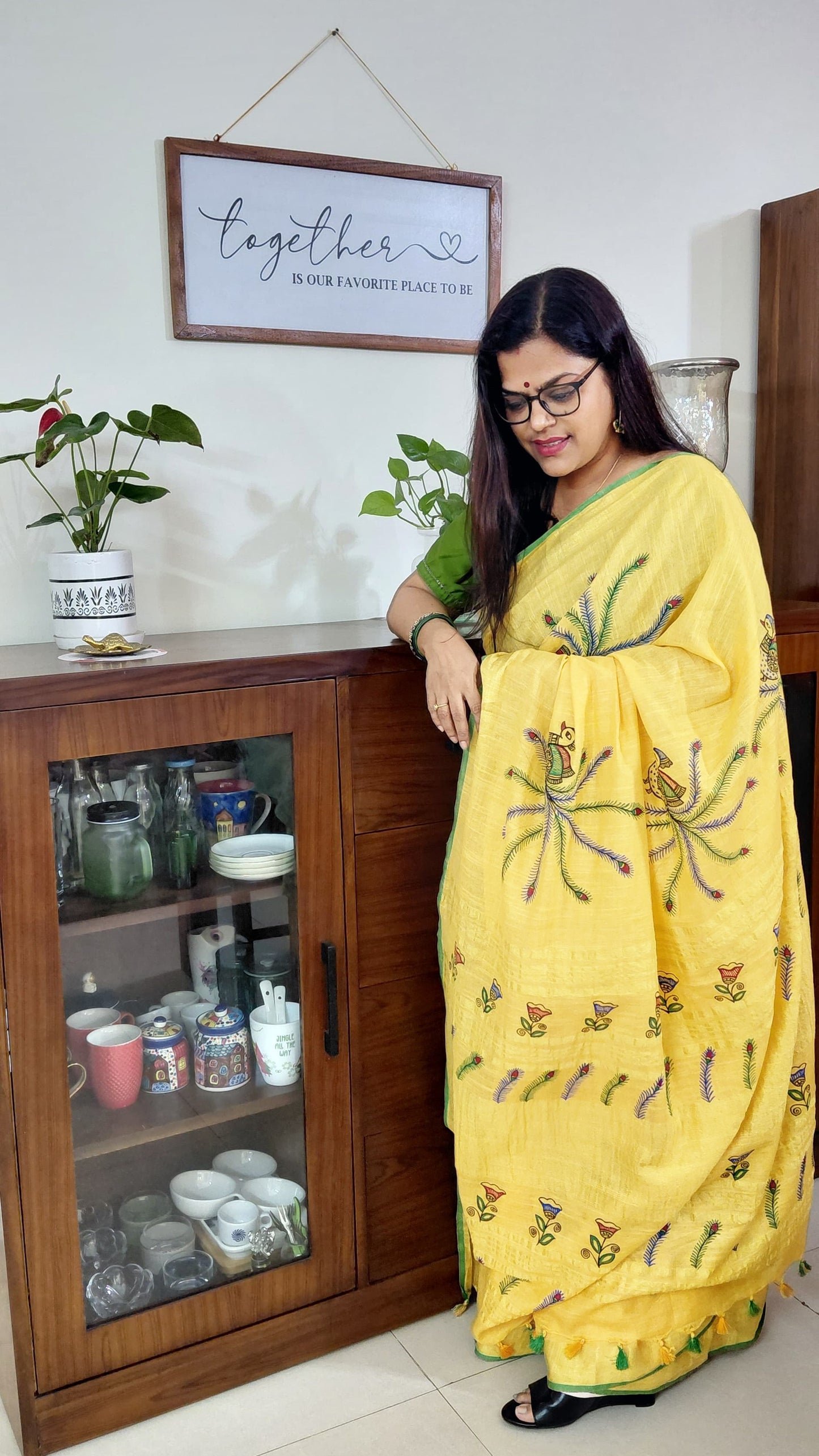 Woman in a yellow saree with floral patterns standing in a room with wooden cabinets and decor.