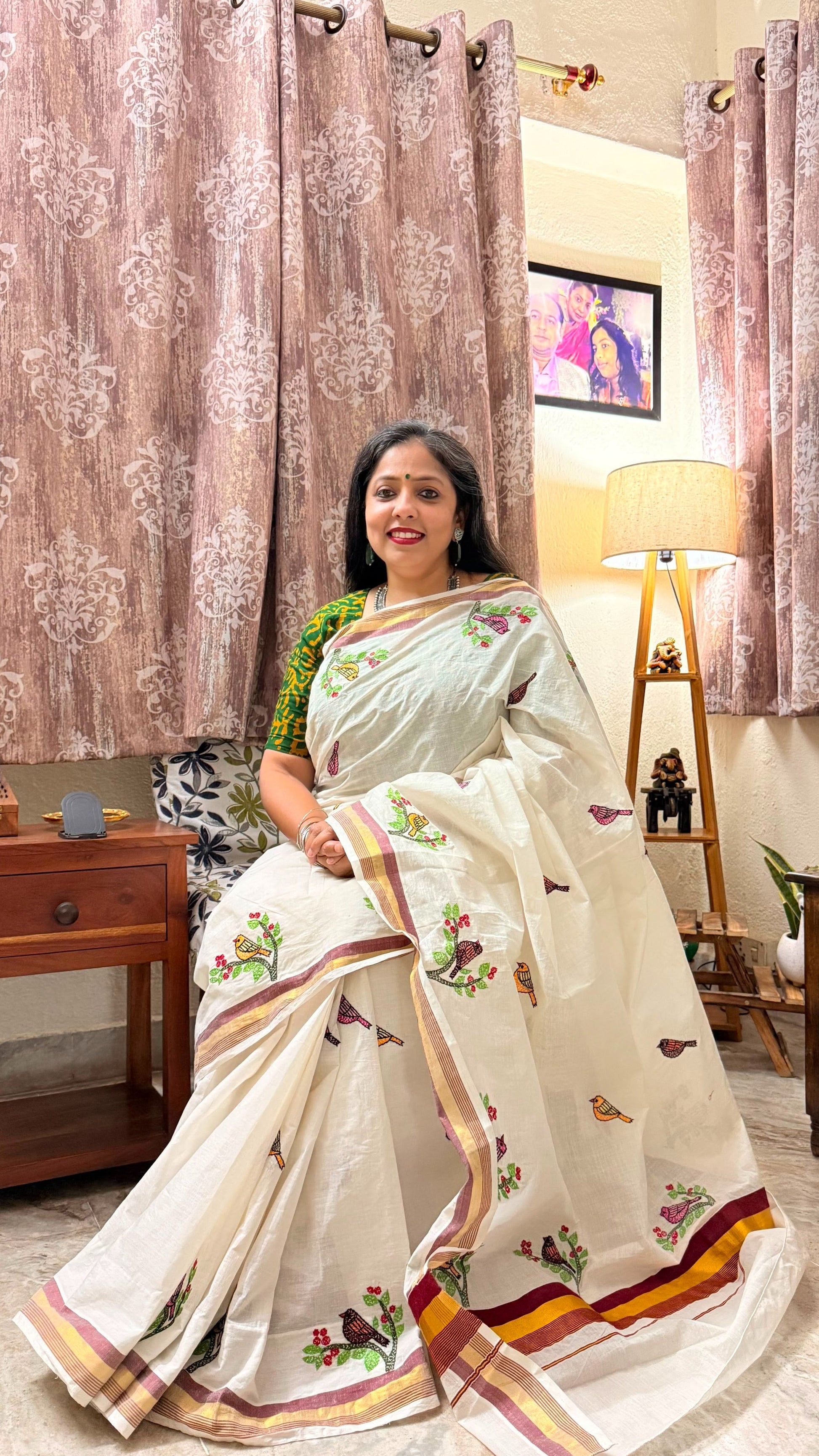 Woman in a Off white Kerala Cotton saree with Sujni Embroidery in a room with decorative curtains and furniture.