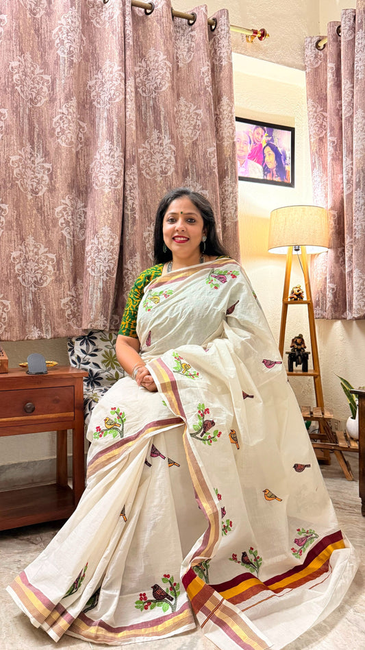 Woman in a Off white Kerala Cotton saree with Sujni Embroidery in a room with decorative curtains and furniture.