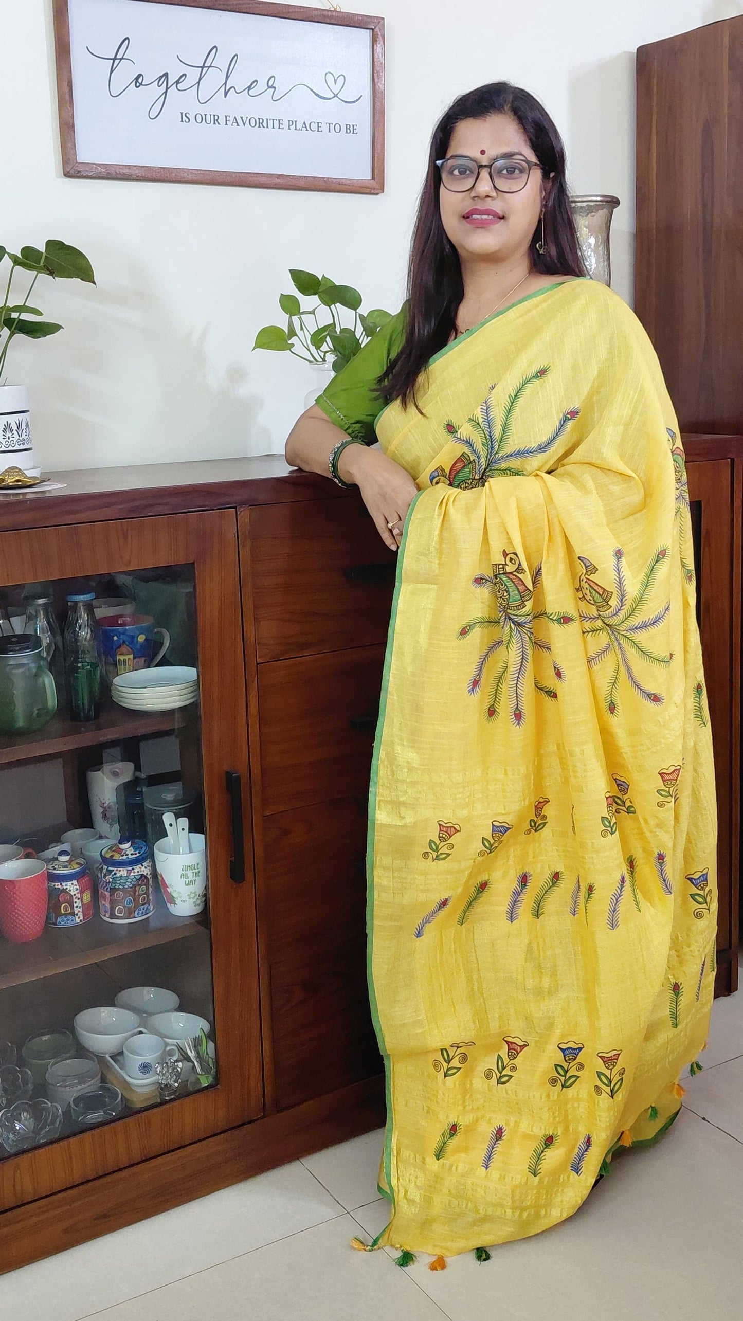 Woman in a yellow linen madhubani saree with floral patterns standing in a room with wooden furniture and decor.