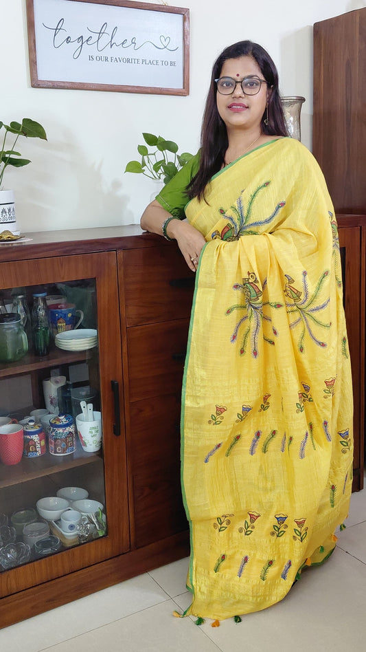 Woman in a yellow linen madhubani saree with floral patterns standing in a room with wooden furniture and decor.
